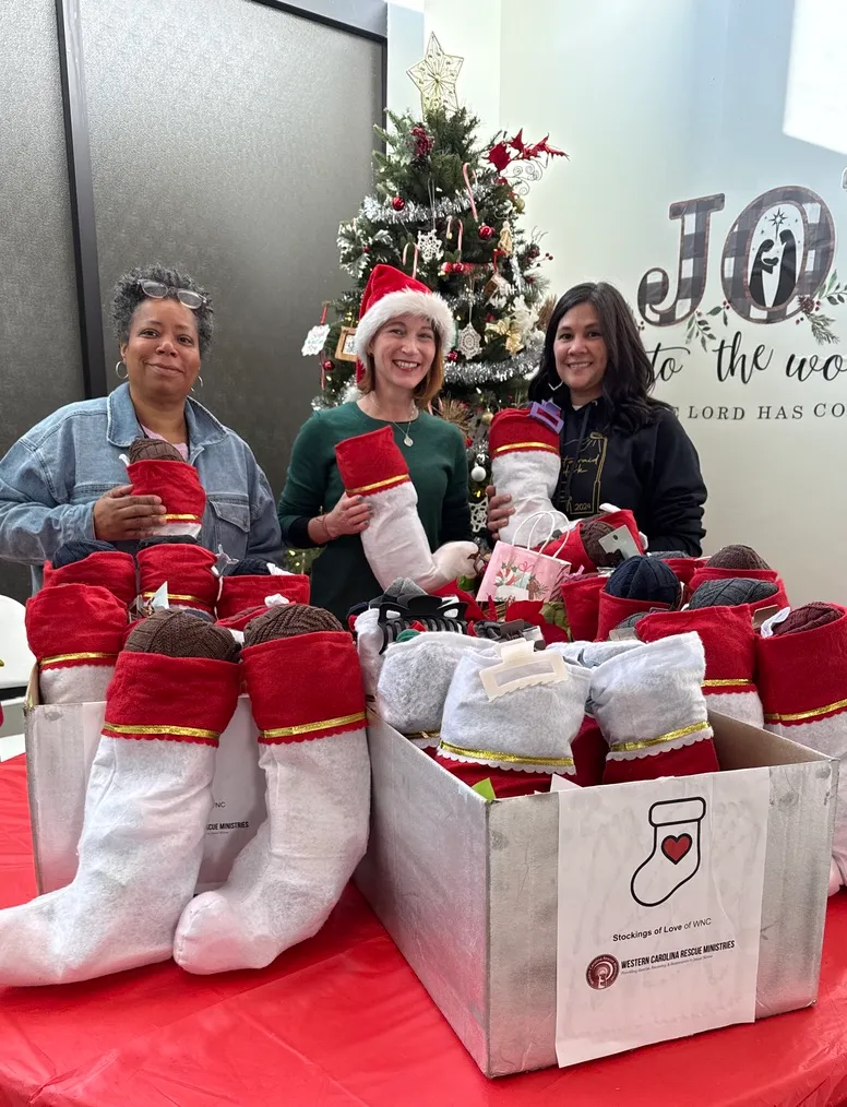 Three women with boxes of filled stockings beside a Christmas tree at Western Carolina Rescue Ministries
